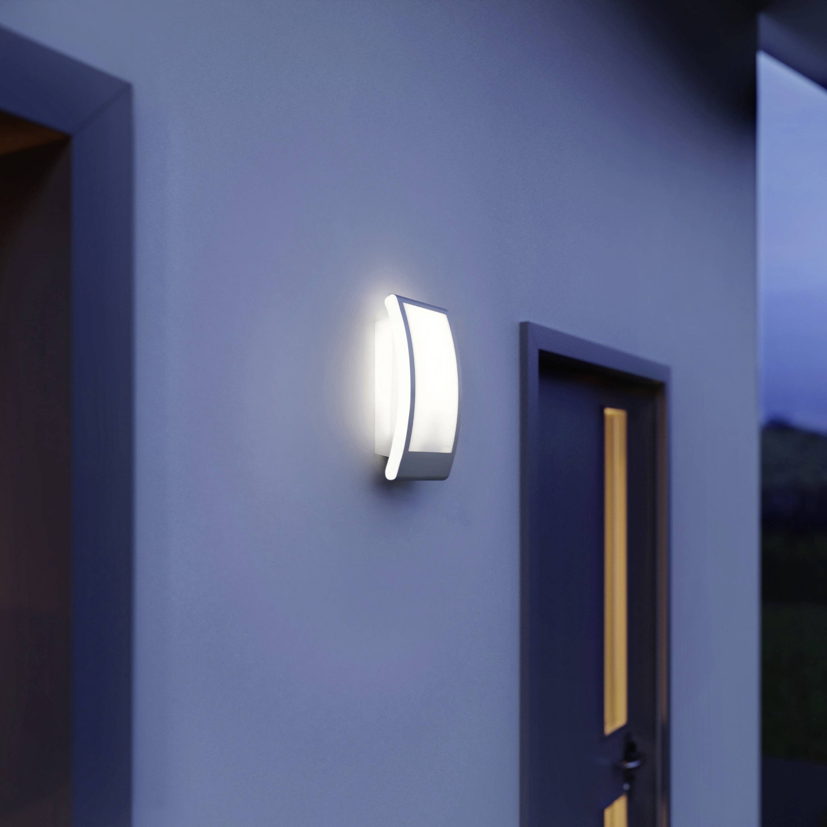 Un luminaire mural moderne diffuse une lumière douce sur un mur bleu, au-dessus d'une porte, illuminant l'entrée au crépuscule.