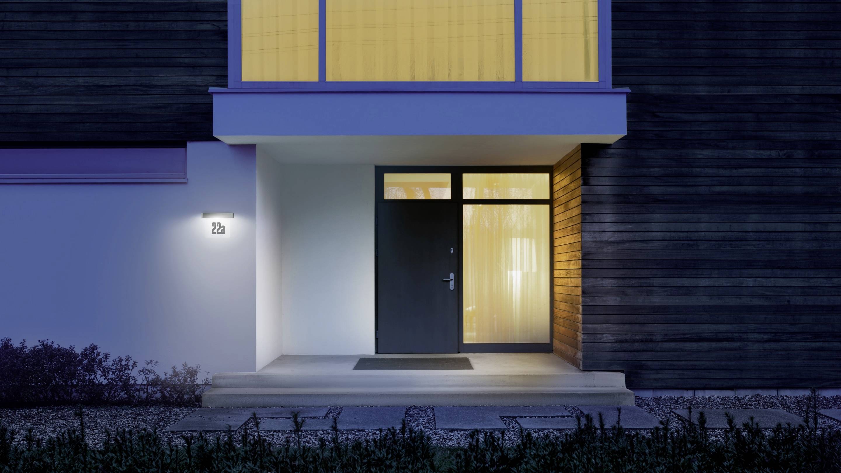 Entrée d'une maison moderne la nuit, éclairée par une lumière chaleureuse venant de l'intérieur. Façade sombre avec de grandes fenêtres.