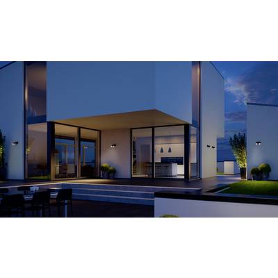 Maison moderne, éclairée au crépuscule avec de grandes fenêtres vitrées montrant une cuisine et un espace de vie. Mobilier de jardin au premier plan.