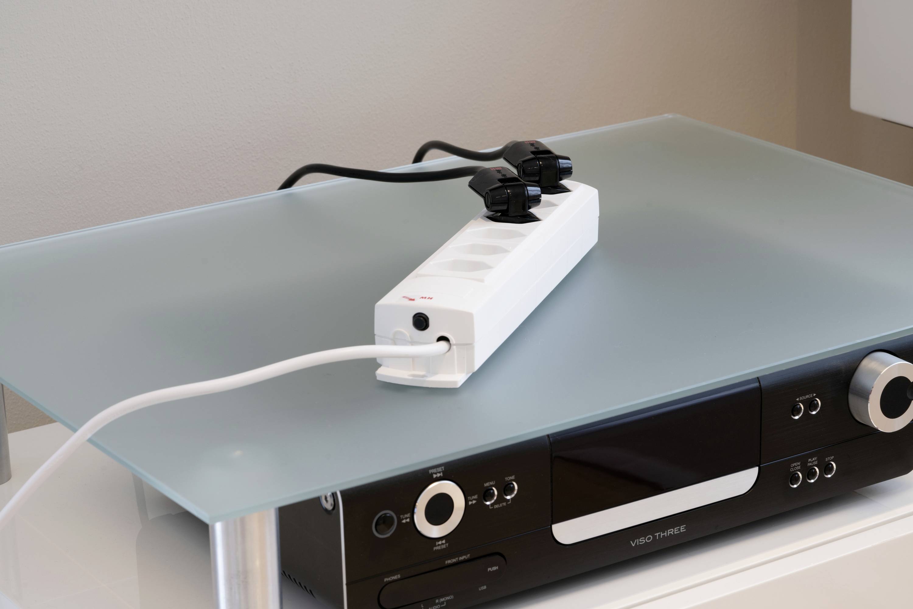 Sur une table en verre se trouve une multiprise blanche avec plusieurs emplacements, sur laquelle deux fiches noires sont branchées. En dessous se trouve un appareil audio.