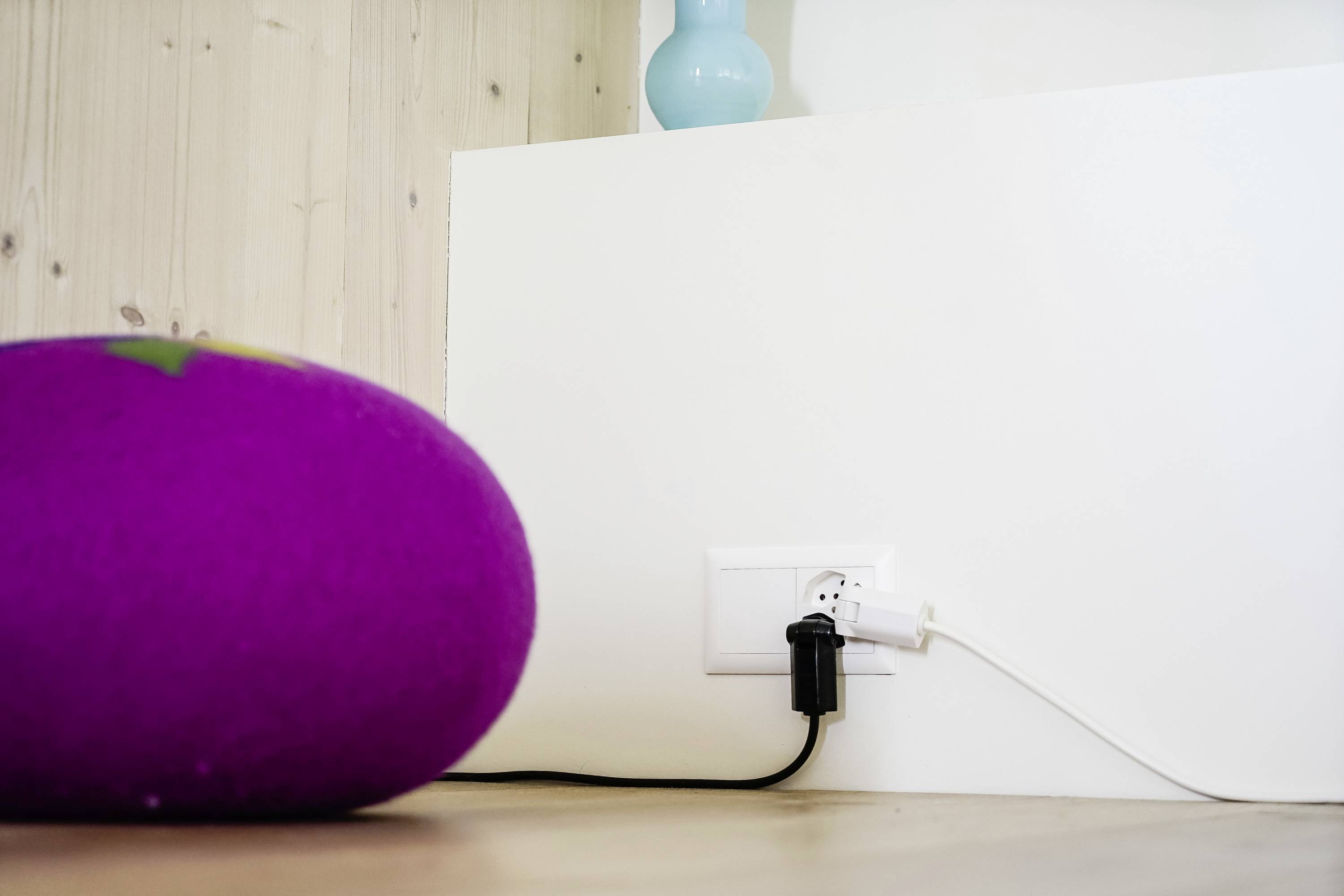 Une chambre blanche avec une prise électrique au mur. Un câble noir et un câble blanc sont branchés. Un coussin violet est posé sur le sol.