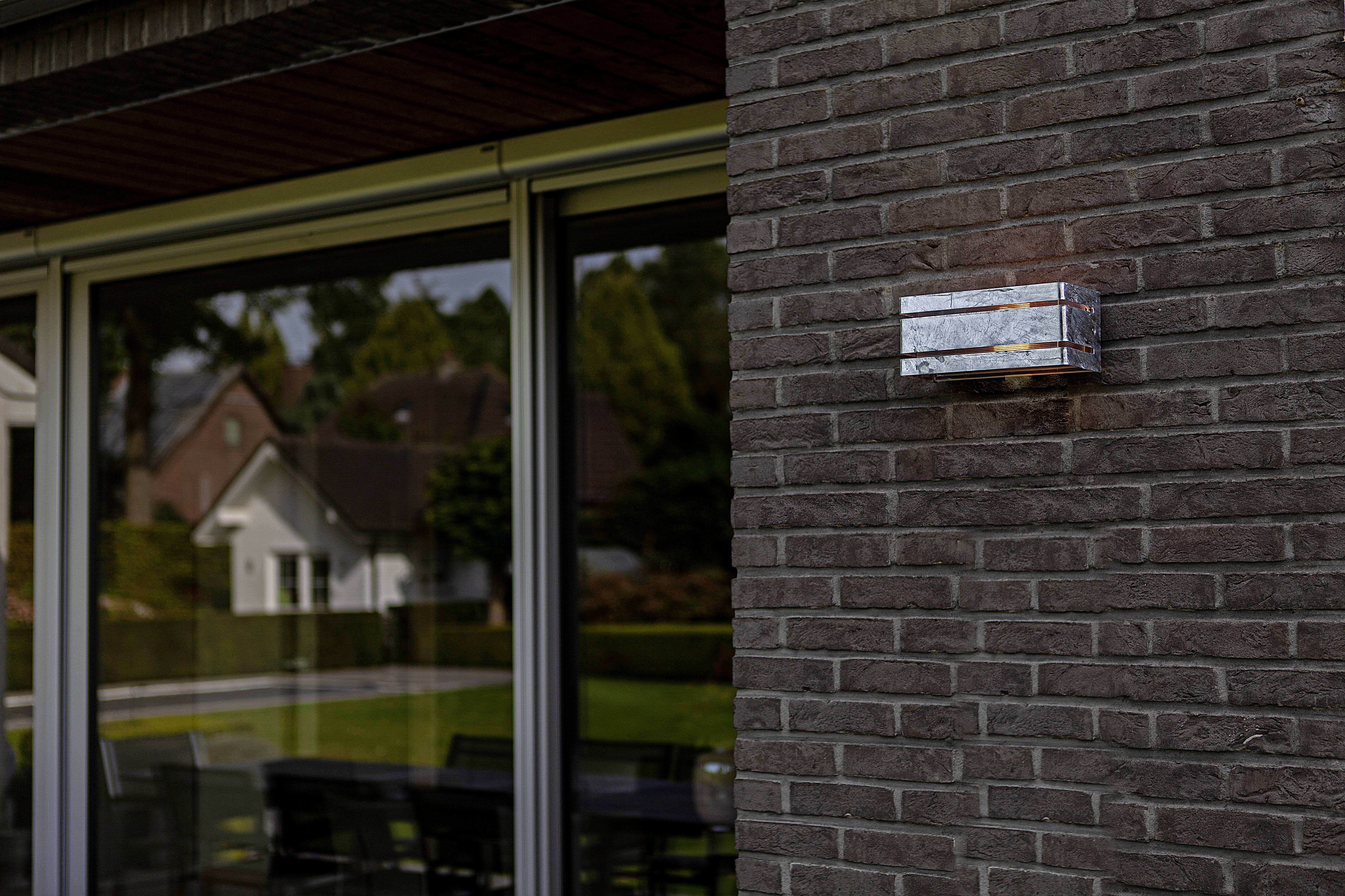 Un luminaire moderne est fixé sur un mur de briques à côté de grandes fenêtres donnant sur un espace de vie avec vue sur un jardin.