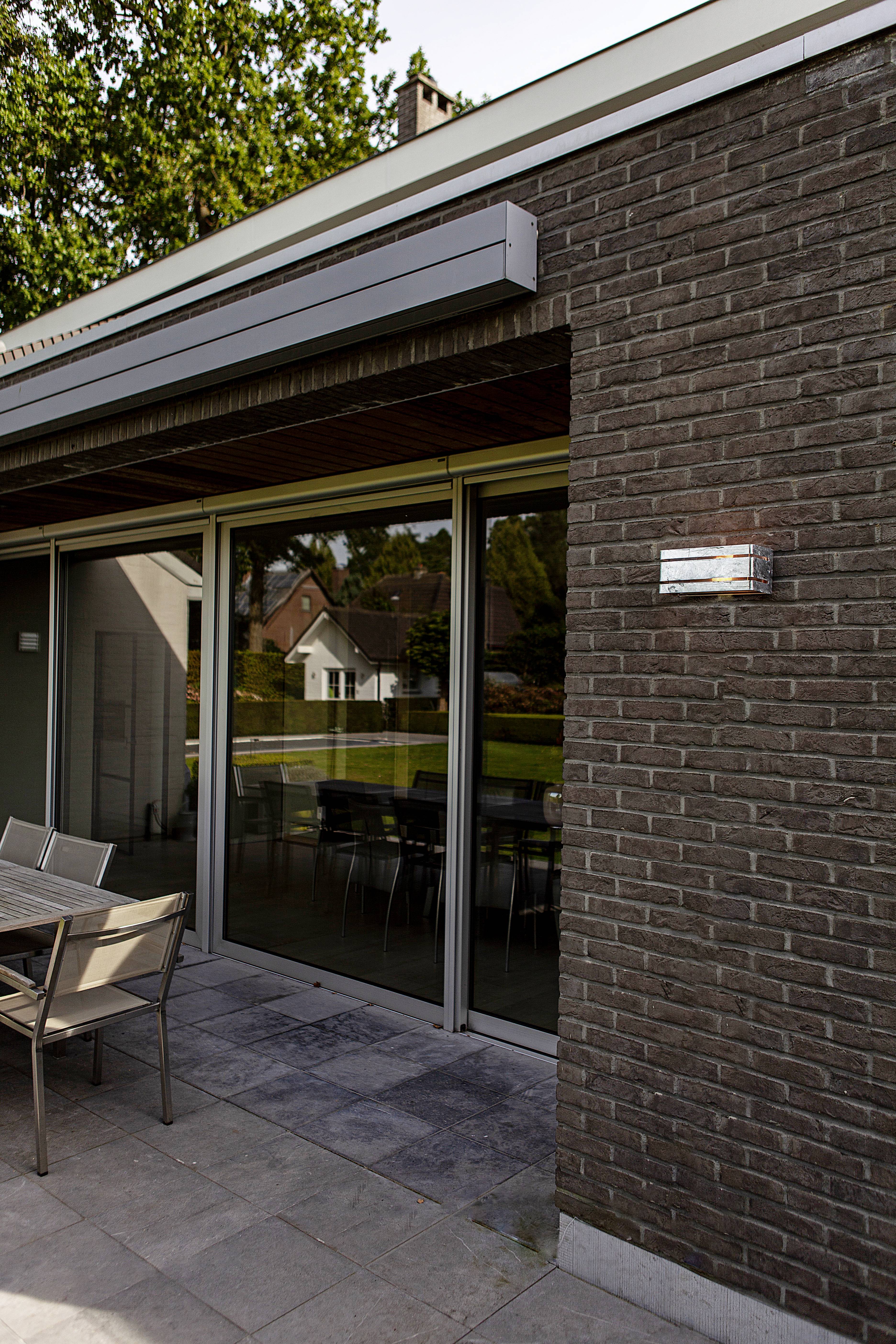 Un bâtiment moderne avec une façade en briques sombres et de grandes portes coulissantes en verre. Devant, une table et des chaises sont disposées sur une terrasse.
