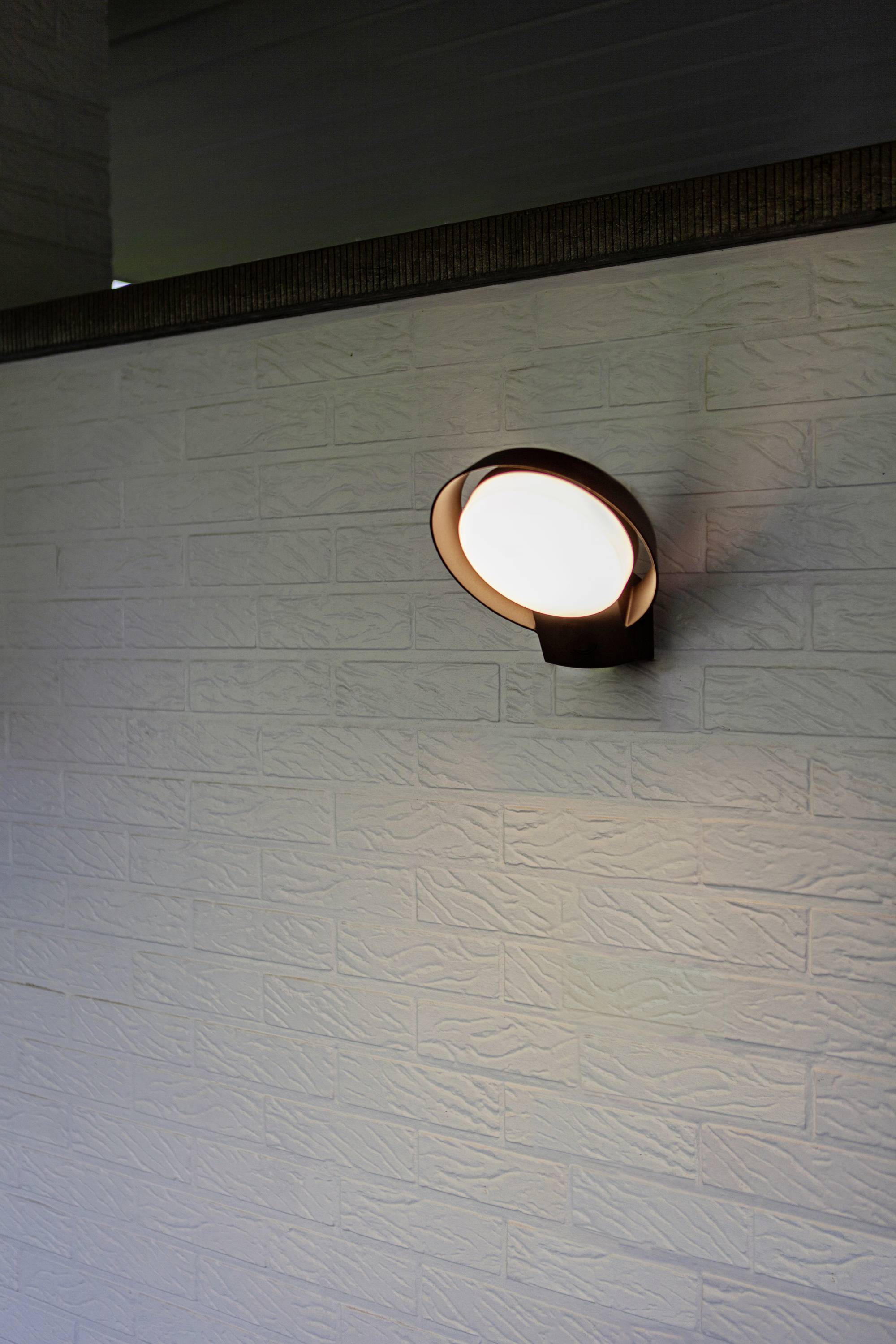 Un applique murale circulaire illumine un mur de briques blanches. La lumière crée une atmosphère douce et chaleureuse dans la pièce.