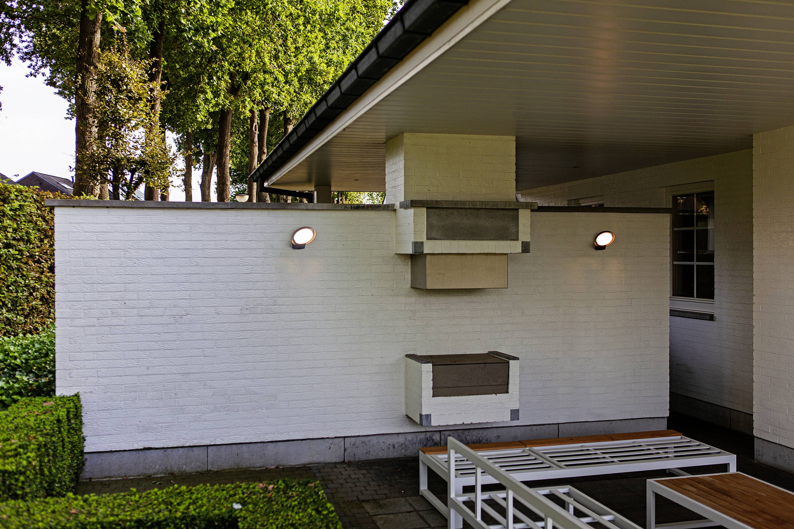 Un grill blanc et moderne est intégré dans un mur de terrasse couvert, entouré de haies et d'arbres, avec des meubles de salon disposés devant.