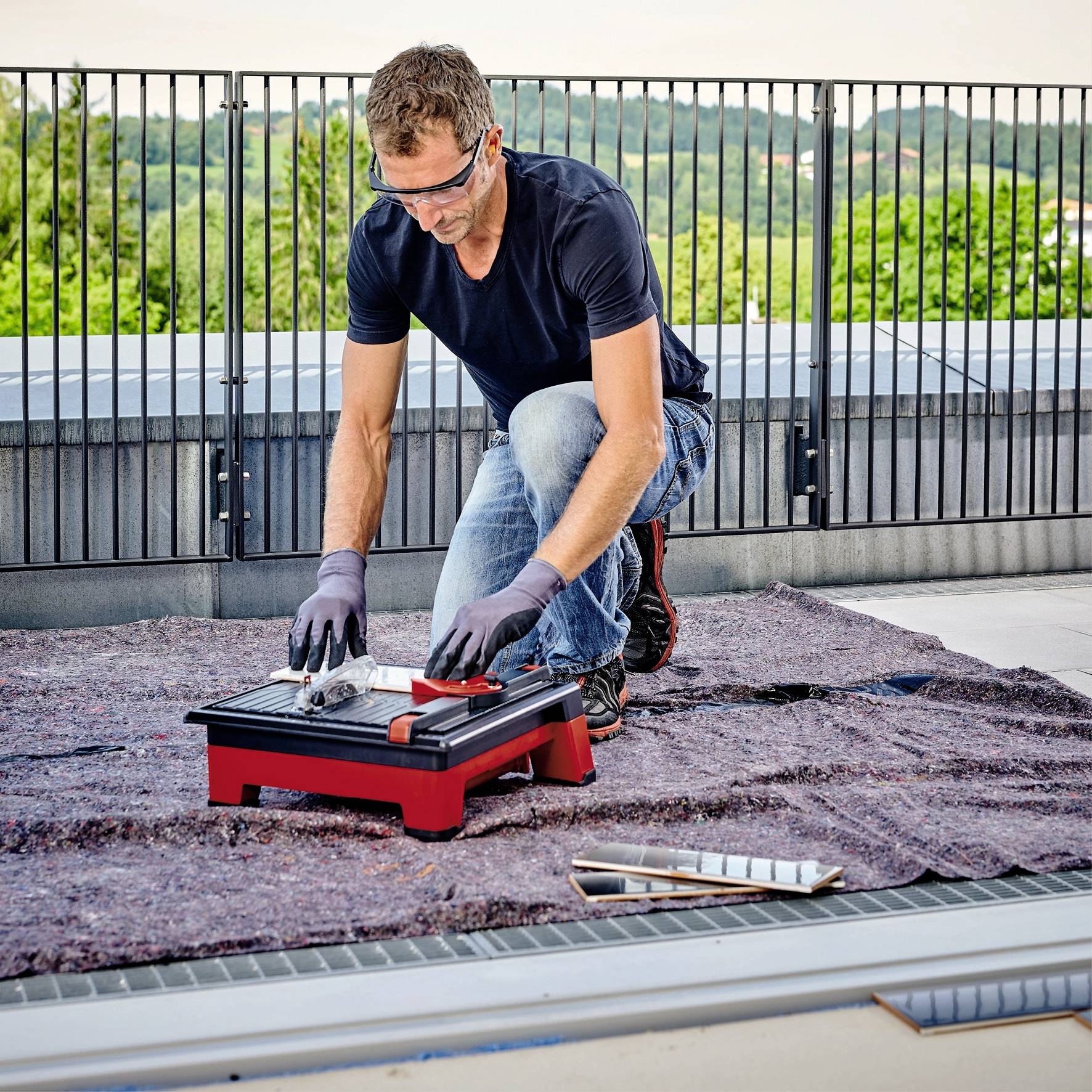 Une personne est agenouillée sur une terrasse et travaille avec concentration sur une petite machine à carrelage, entourée d'autres outils.