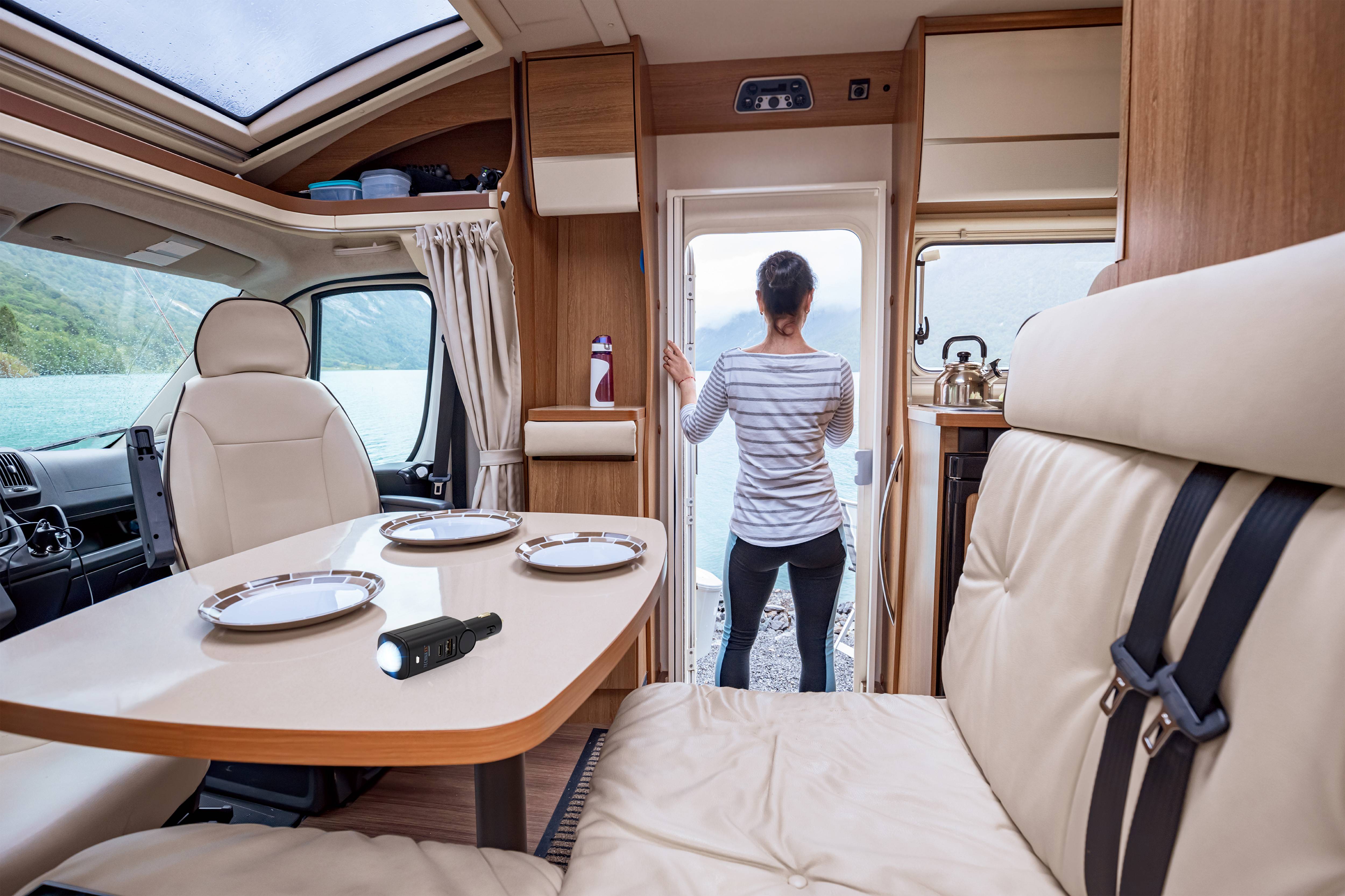Intérieur d'un camping-car avec une table à manger et une assiette. Une personne se tient près de la porte ouverte, regardant un lac et des montagnes.