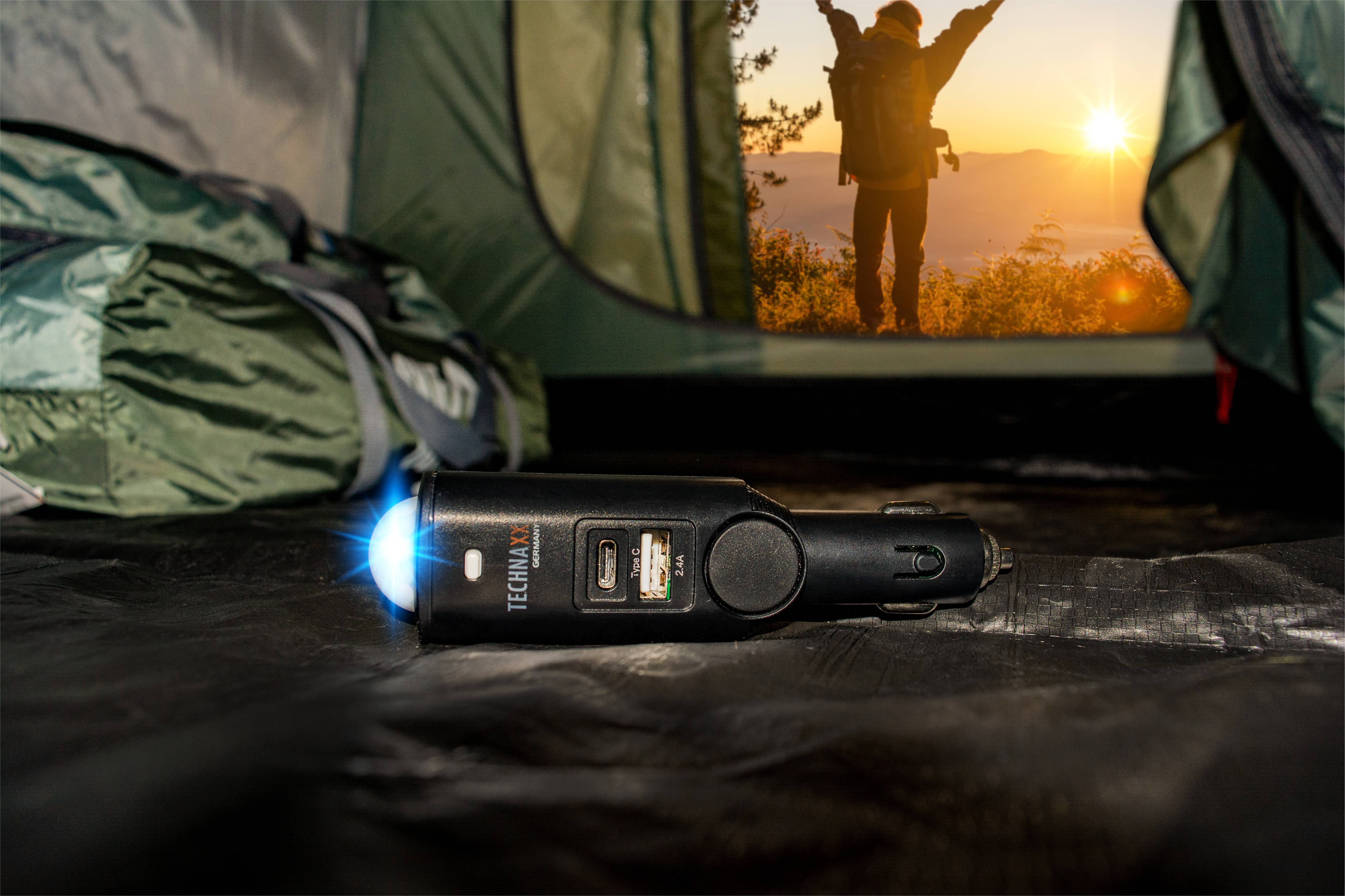 Un câble de charge est allongé dans une tente illuminée, avec en arrière-plan une personne qui lève les bras dans la nature au lever du soleil.