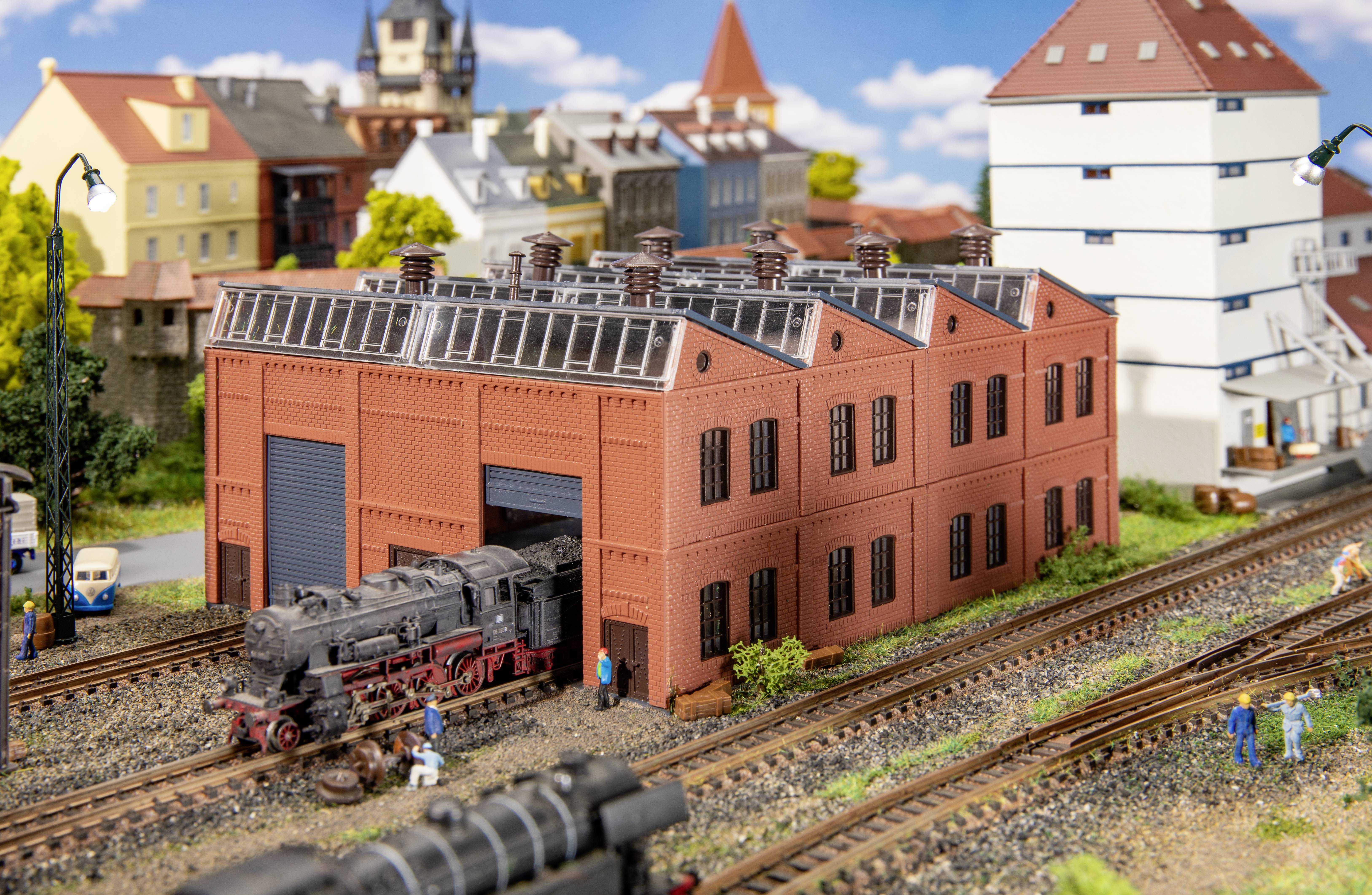 Un diorama de train miniature montre une locomotive à vapeur noire sortant d'un bâtiment en briques rouges. En arrière-plan, on aperçoit des bâtiments et des arbres.