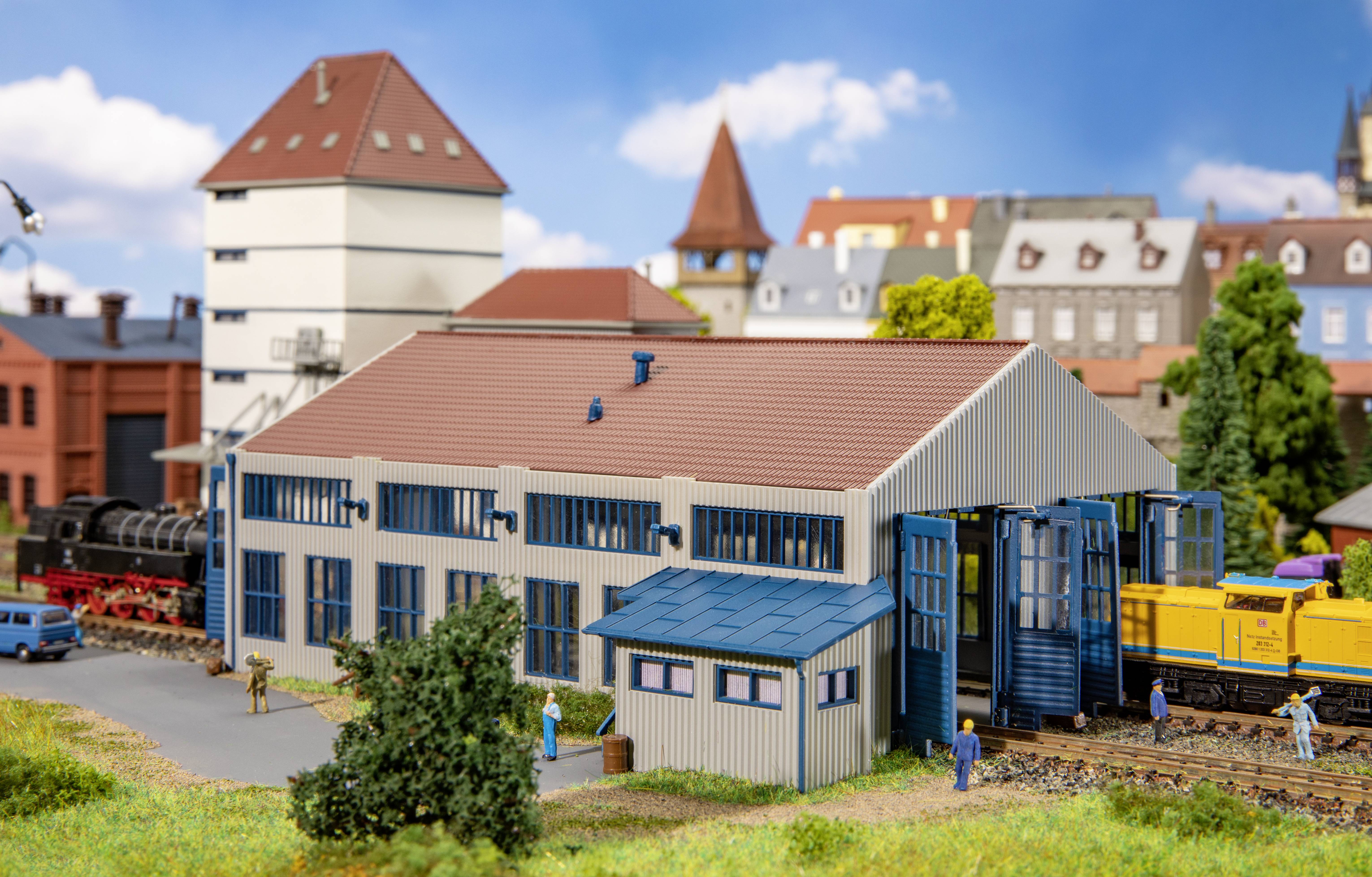 Maquette miniature d'un hangar ferroviaire avec sa porte ouverte, entouré de figurines et d'arbres, avec un décor de ville en arrière-plan.