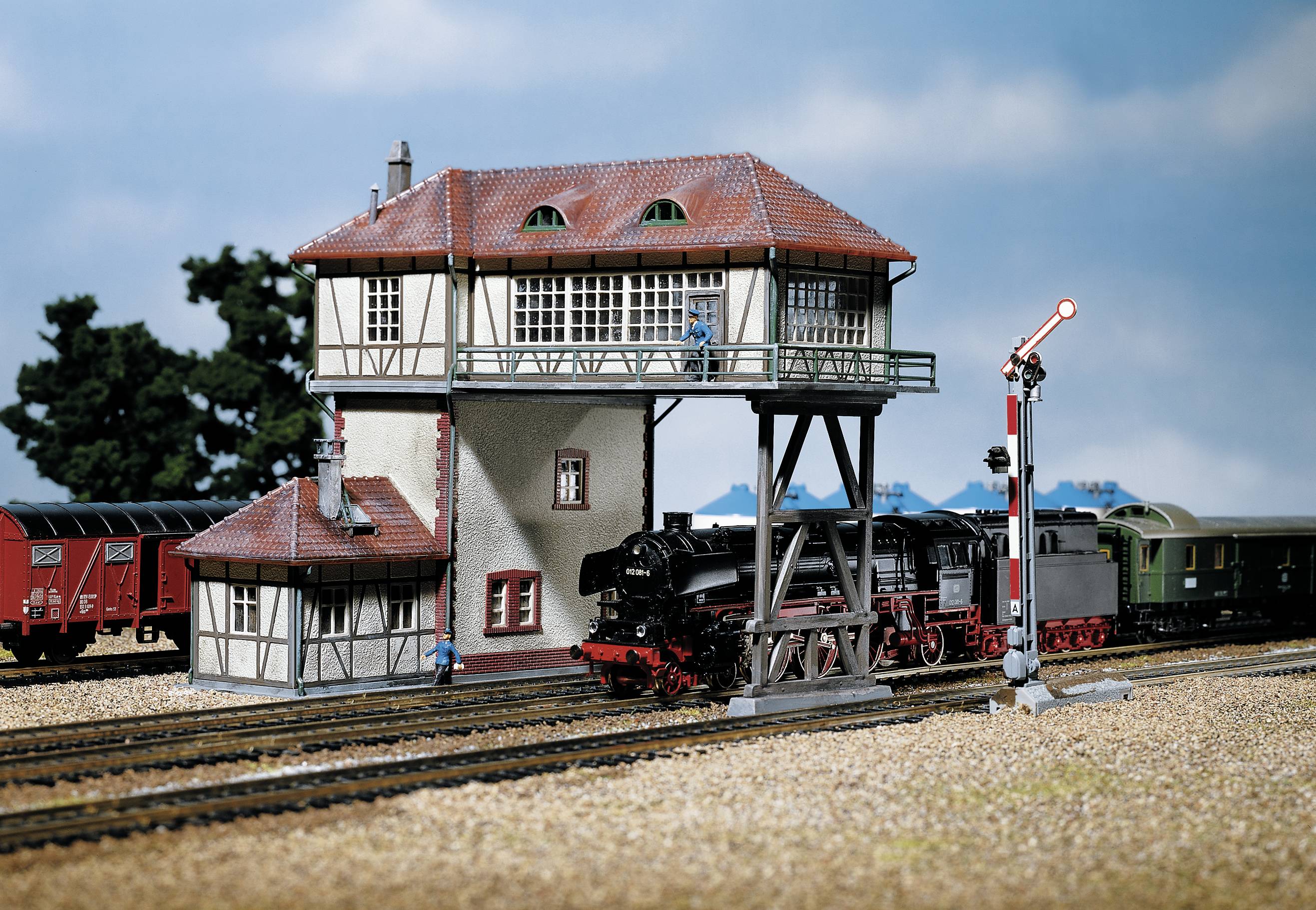 Une locomotive à vapeur roule devant une maisonnette de garde-barrière détaillée sur un terrain de modélisme ferroviaire, accompagnée de wagons et de signaux en arrière-plan.