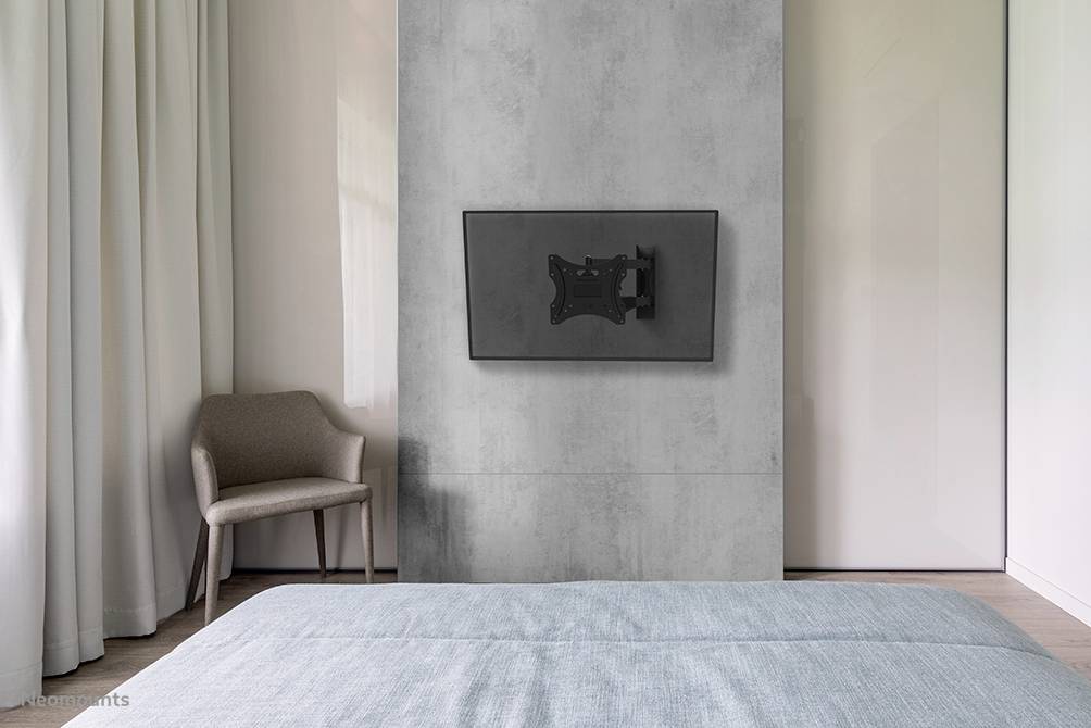 Une chambre moderne avec un fauteuil gris, un lit recouvert d'un jeté bleu et une télévision montée au mur.