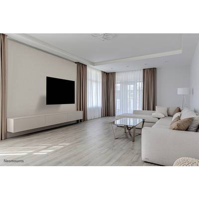 Grand salon avec un parquet clair, un canapé beige avec des coussins, une table basse en verre, une télévision montée au mur et de grandes fenêtres.