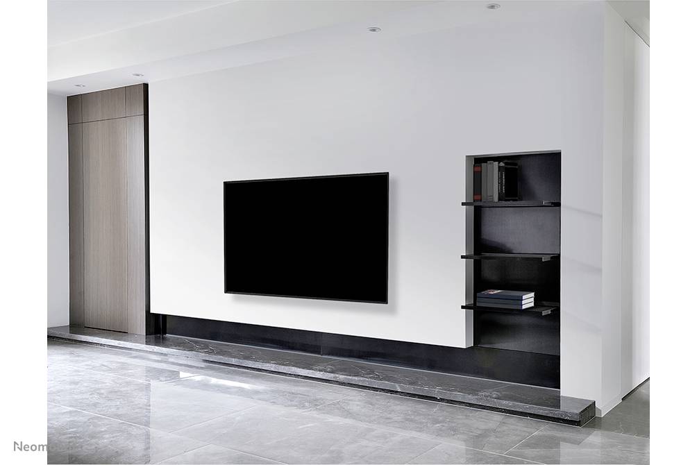 Un salon avec un grand téléviseur noir sur le mur blanc. À côté, des étagères flottantes garnies de livres et de décorations.
