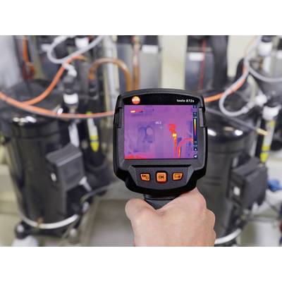Une main tient une caméra thermique qui capture des équipements techniques en arrière-plan. L'écran affiche des répartitions de température.