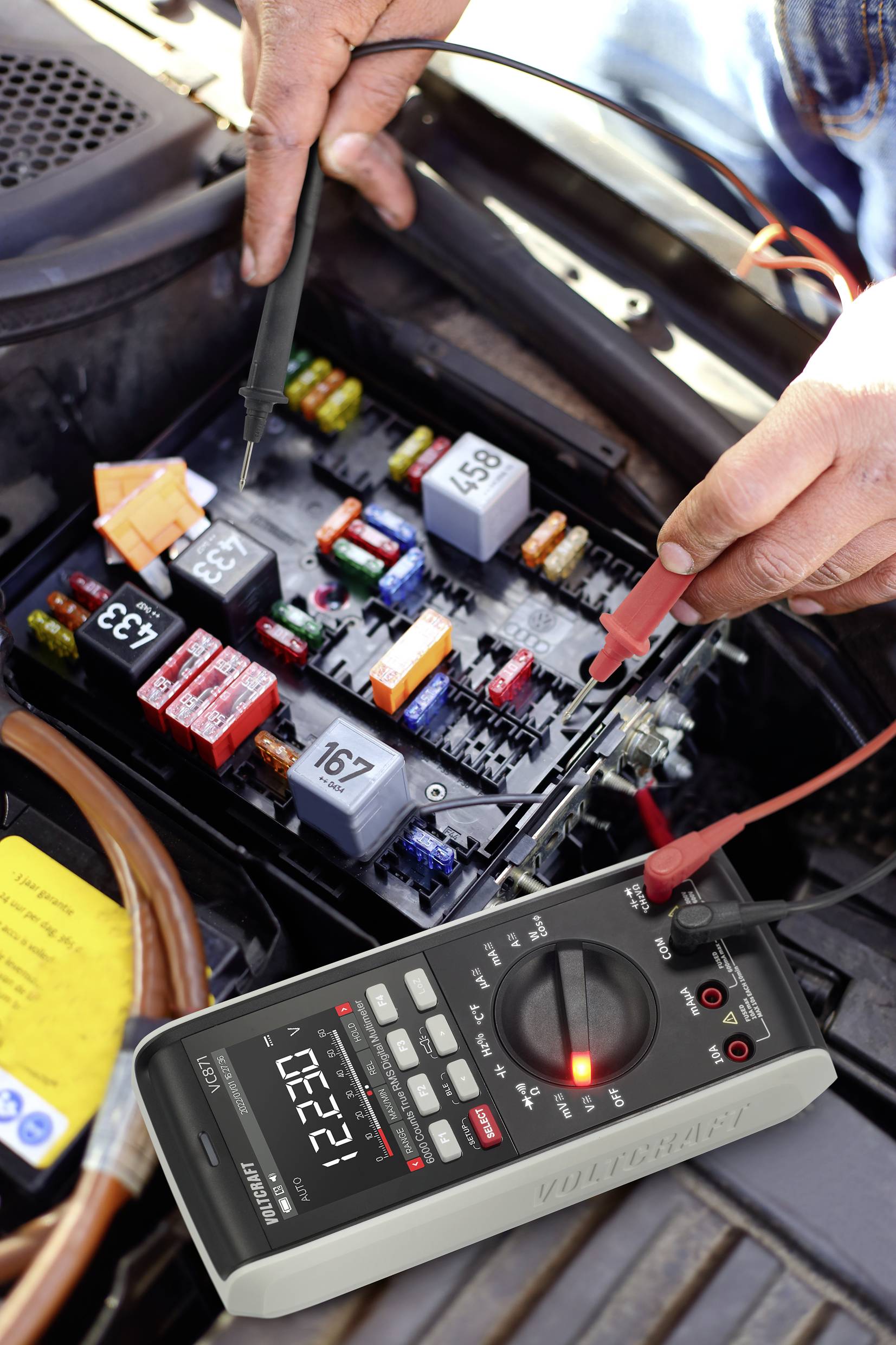 Une personne mesure la tension dans un boîtier de fusibles automobile à l'aide d'un multimètre. La mesure indique 12,27 volts.