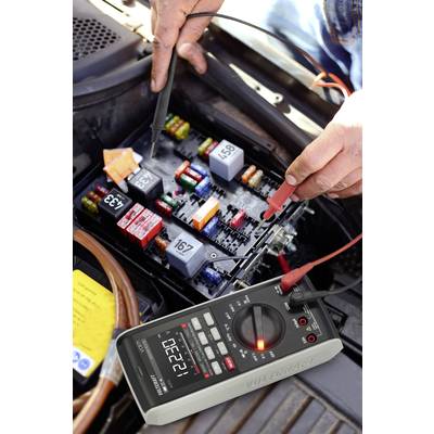 Une personne mesure la tension dans un boîtier de fusibles automobile à l'aide d'un multimètre. La mesure indique 12,27 volts.