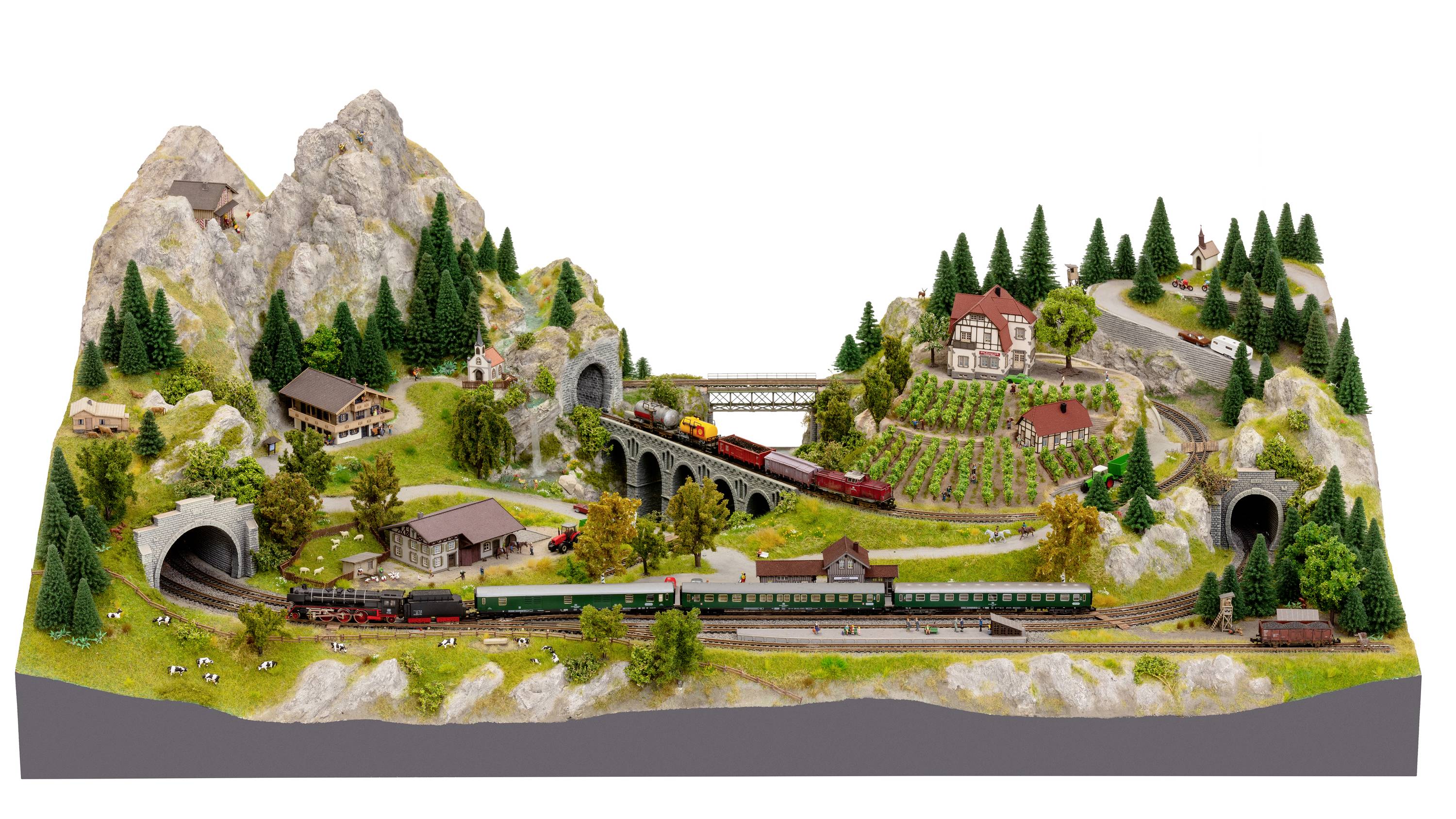 Un paysage ferroviaire miniature détaillé avec des montagnes, des tunnels, des trains et des bâtiments. Un train circule sur un pont surélevé.