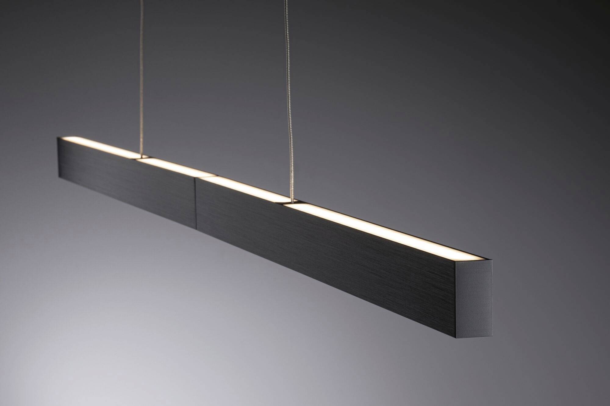 Une lampe suspendue moderne et linéaire en métal sombre flotte devant un fond gris et diffuse une lumière uniforme.