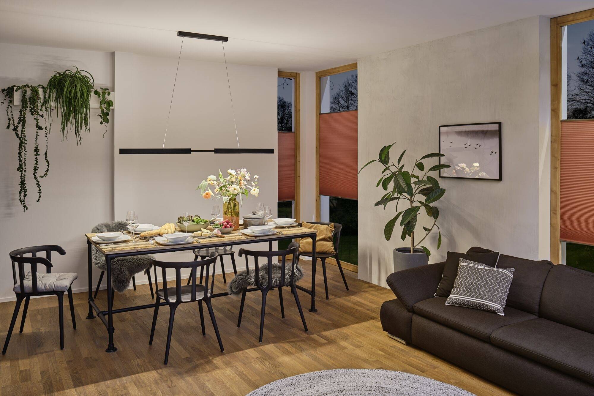 Salle à manger avec table et chaises, décorée avec des fleurs dans un vase. Lampe suspendue moderne et plantes vertes en arrière-plan.