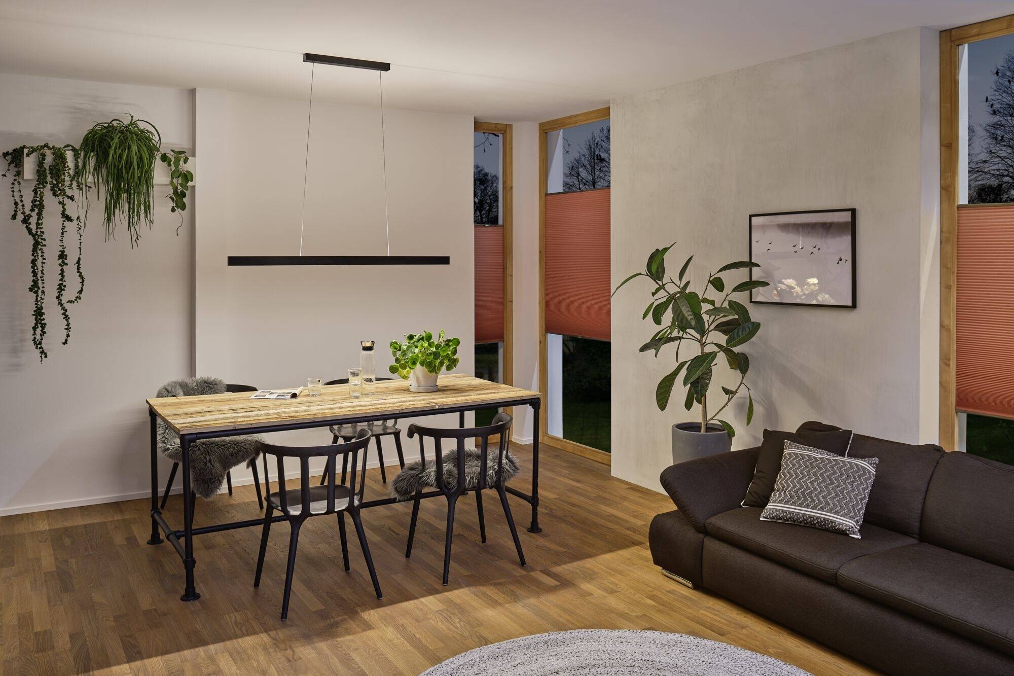 Salle à manger avec une table en bois et quatre chaises, des plantes, une suspension, un canapé à droite. Fenêtre avec des stores roses, ambiance du soir à l'extérieur.