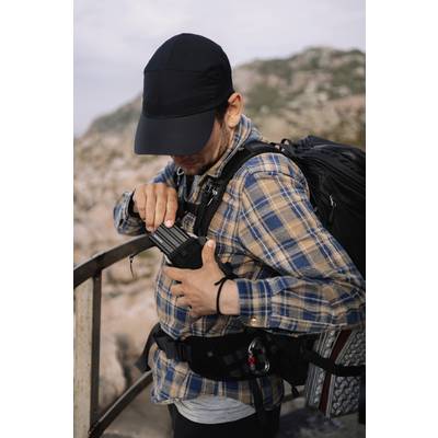 Une personne se tient sur une rambarde dans un paysage montagneux, portant des vêtements à carreaux, un sac à dos et manipulant un appareil.