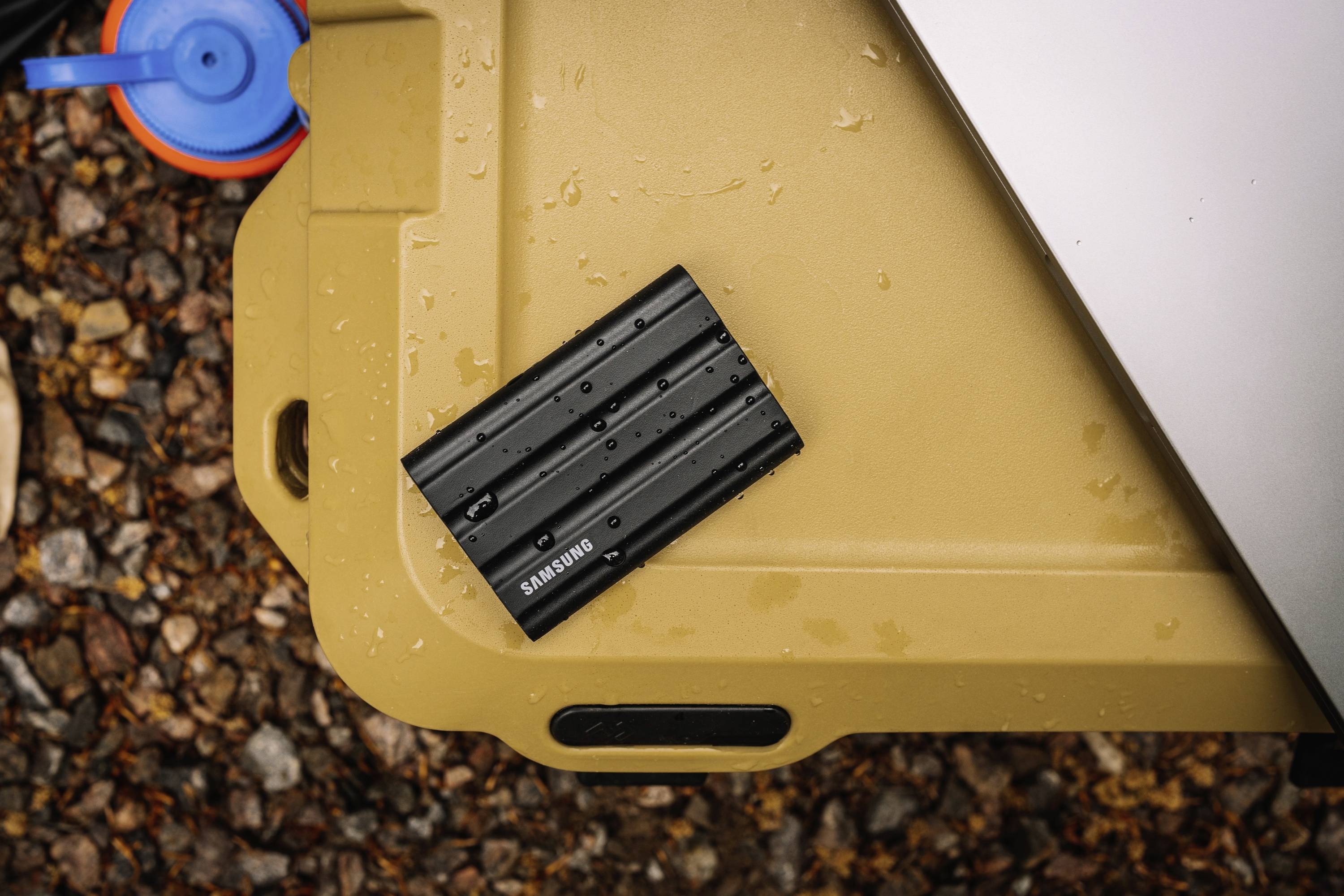 Un disque dur externe noir, résistant à l'eau, de la marque 'Samsung' est posé sur un fond beige à l'extérieur.