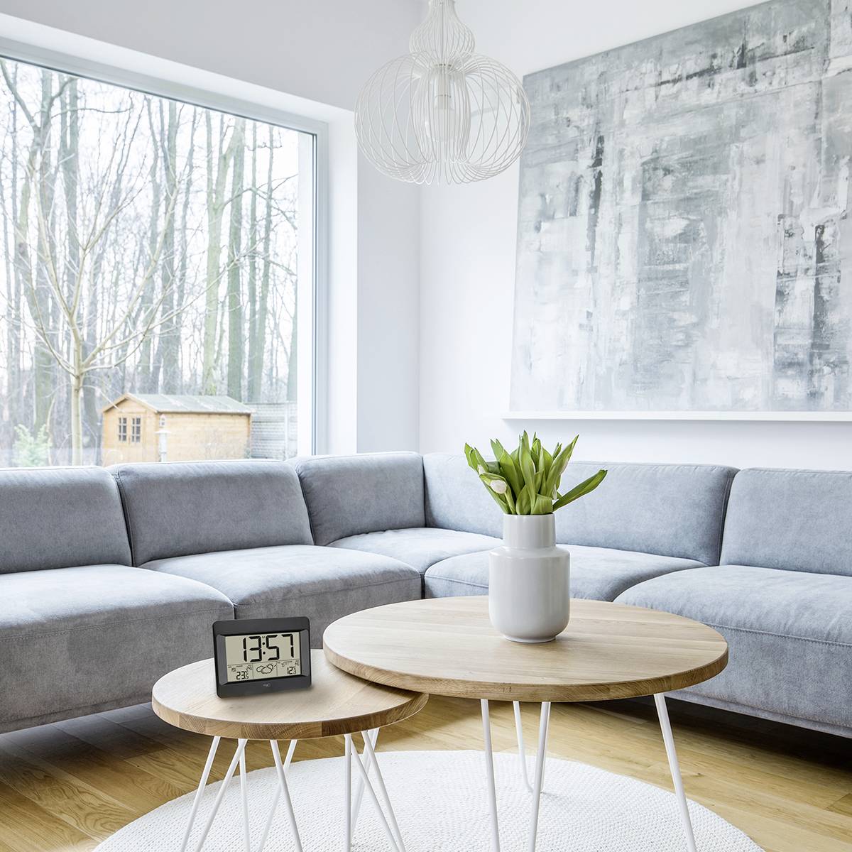 Salon lumineux avec un canapé gris, une table ronde en bois et une horloge numérique. Grande œuvre d'art murale et fenêtre donnant sur des arbres.
