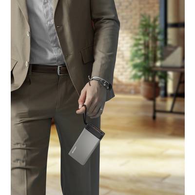 Une personne en costume tient un disque dur externe à la main. À l'arrière-plan, on aperçoit un bureau avec un sol en bois et des plantes en pot.