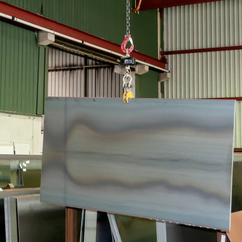 Une plaque d'acier est transportée dans un atelier à l'aide d'un pont roulant. L'environnement présente une architecture industrielle et des étagères de stockage.