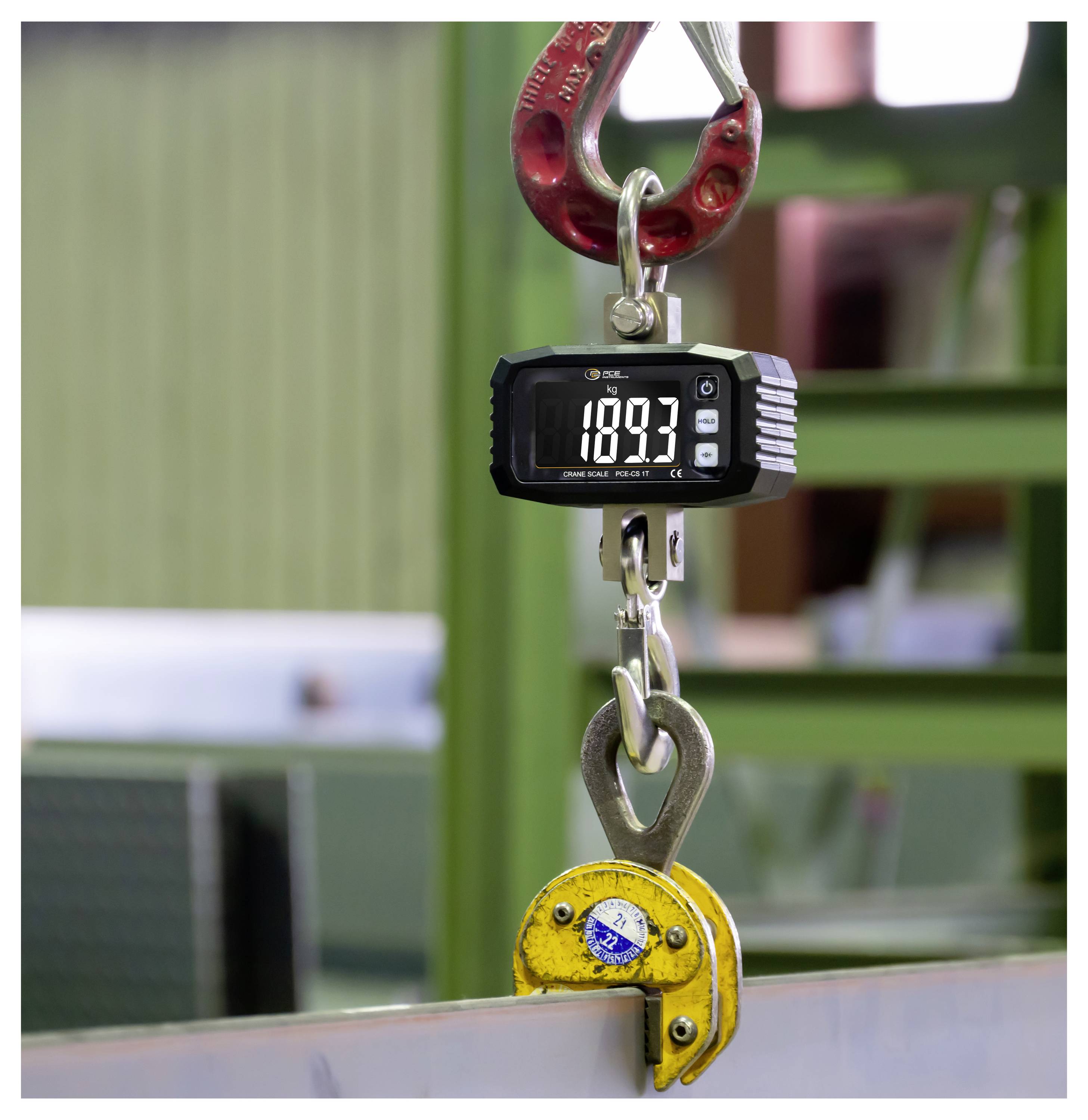Balance numérique suspendue affichant 1893 kg, suspendue à un crochet de grue rouge et fixée à une pince de levage jaune sur une poutre métallique.