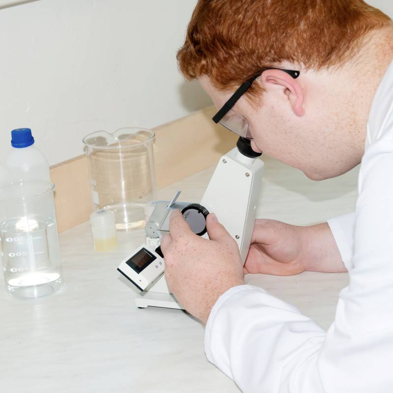 Un homme examine une lame au microscope dans un laboratoire. Plusieurs récipients contenant des liquides sont posés sur la table.
