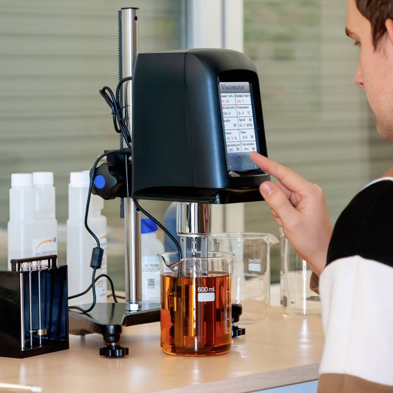 Une personne manipule un viscosimètre dans un laboratoire. Un bécher contenant un liquide est placé sur l'appareil. Des ustensiles de laboratoire sont visibles en arrière-plan.