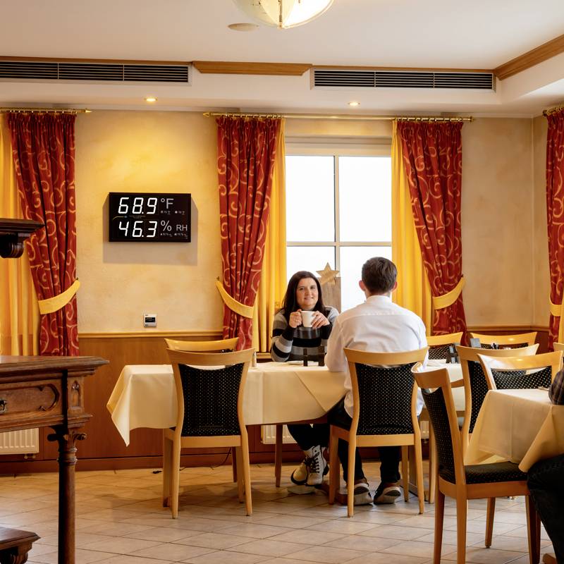 Un couple est assis à une table dans un restaurant, entouré de rideaux jaunes. Un affichage sur le mur indique 20,5°C et 46,3% d'humidité relative.