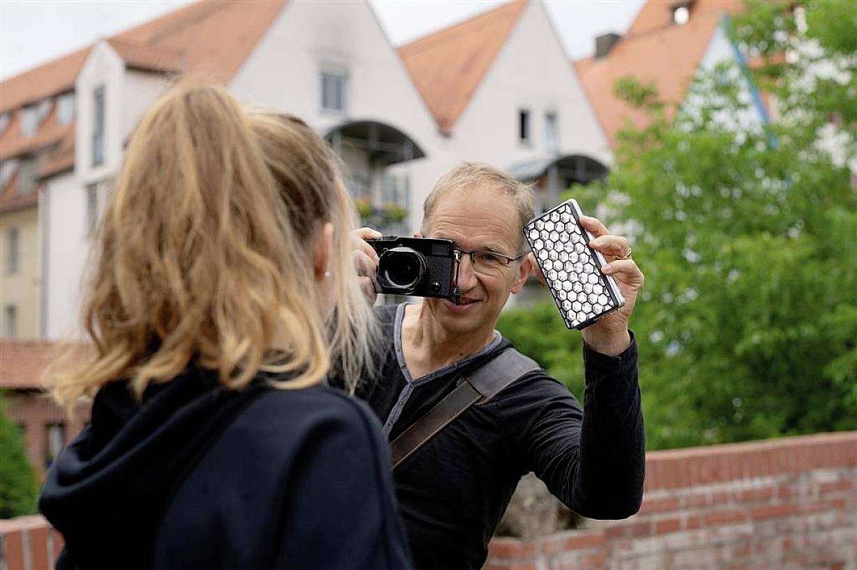 Un homme photographie une femme à l'extérieur tout en tenant un réflecteur. Des maisons sont visibles en arrière-plan.