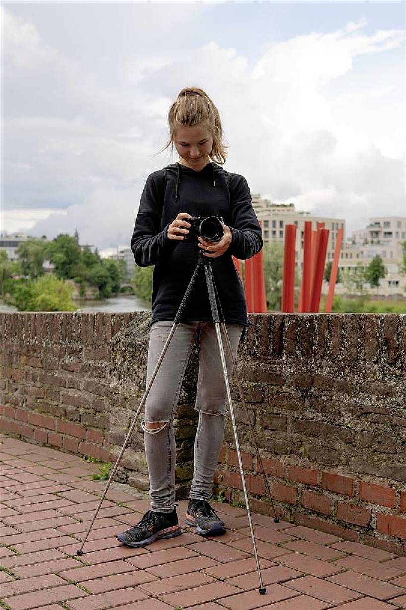 Une personne se tient dehors sur un chemin de briques. Elle tient un appareil photo avec un trépied et regarde vers le bas. Des arbres et des bâtiments sont visibles en arrière-plan.