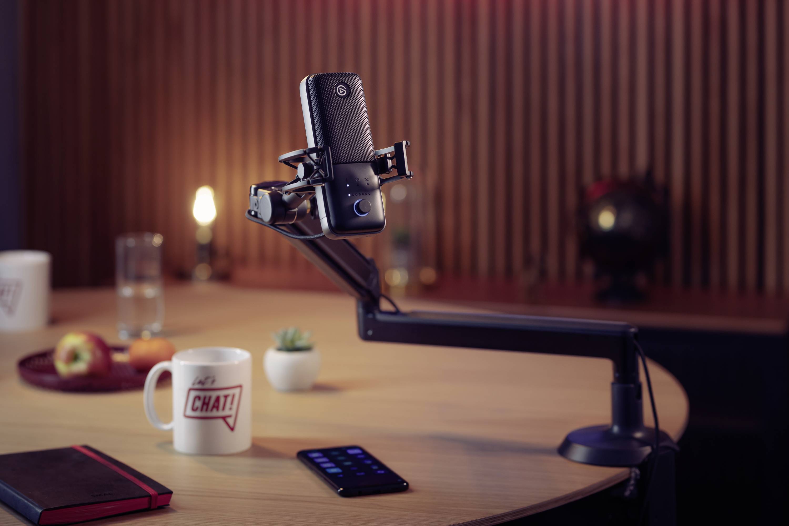 Un smartphone est fixé dans un support sur une table. En arrière-plan, on trouve des tasses, un carnet et des plantes.