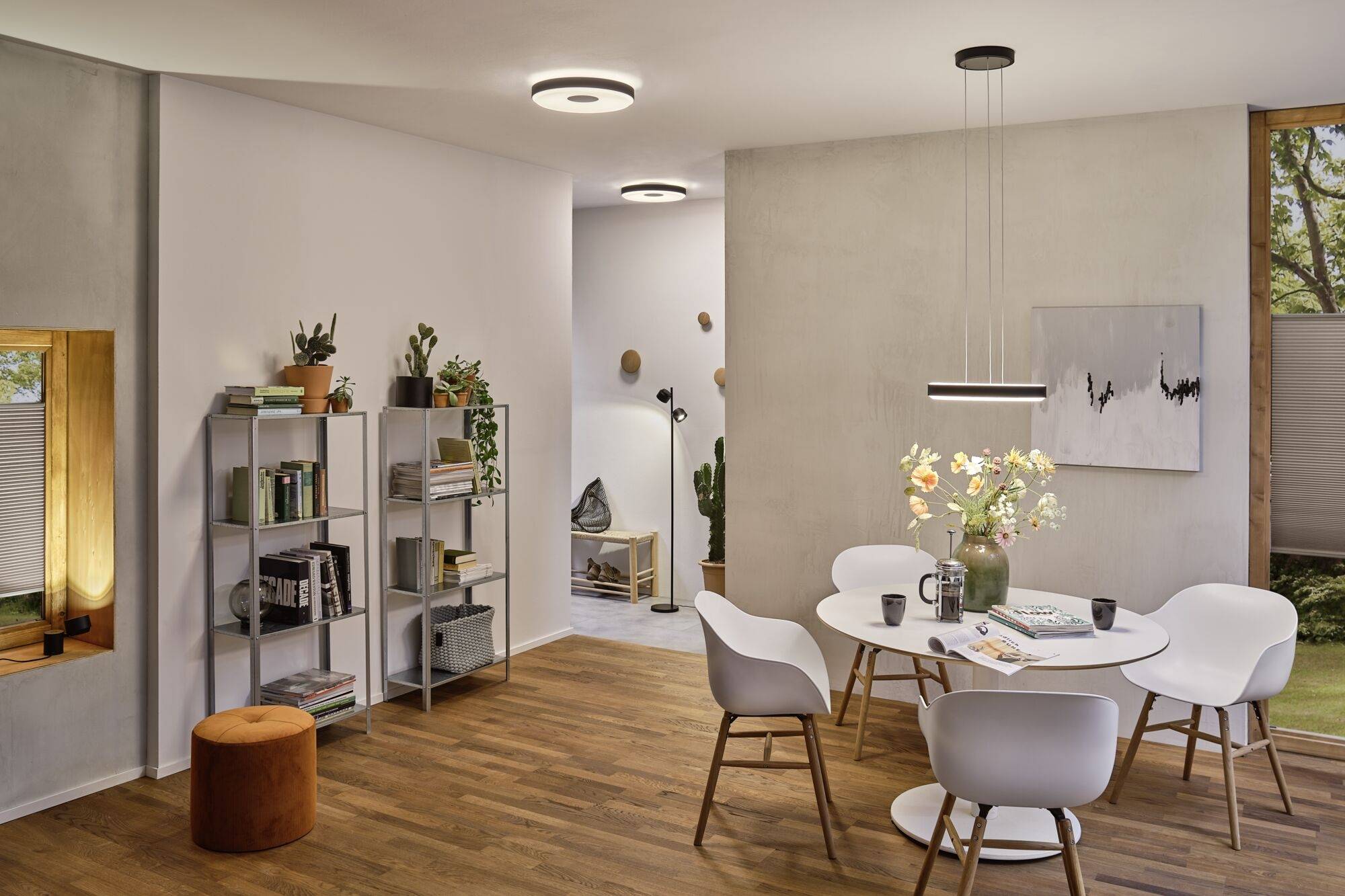 Salle à manger avec une table ronde et cinq chaises blanches, entourée de plantes et d'une bibliothèque, éclairée de manière moderne. La fenêtre donne sur un jardin verdoyant.