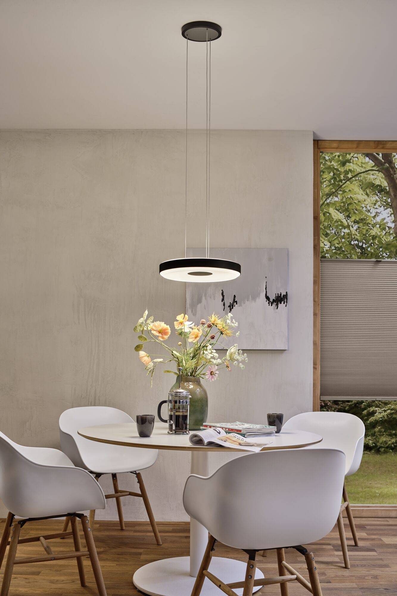 Espace repas moderne avec une table ronde, quatre chaises blanches et un arrangement floral au centre. Une suspension lumineuse éclaire la pièce.
