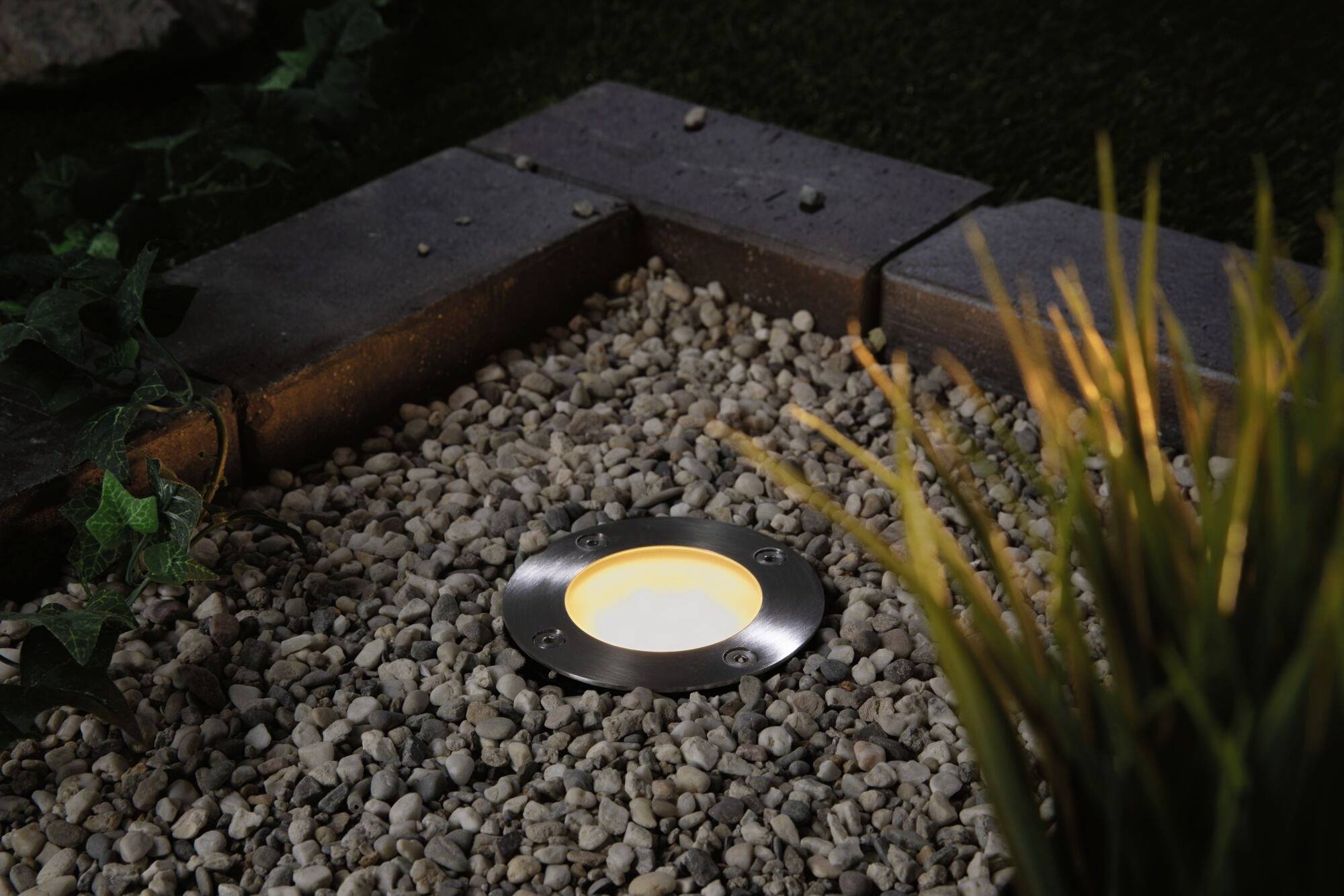 Éclairage au sol dans une zone de graviers près de plantes et de pierres dans un jardin au crépuscule, créant une atmosphère chaleureuse.