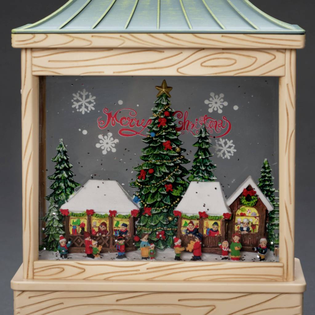 Une scène de Noël en diorama avec des figurines, un sapin et des maisons. En arrière-plan, "Merry Christmas" est écrit devant des flocons de neige.