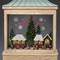 Une scène de Noël en diorama avec des figurines, un sapin et des maisons. En arrière-plan, "Merry Christmas" est écrit devant des flocons de neige.