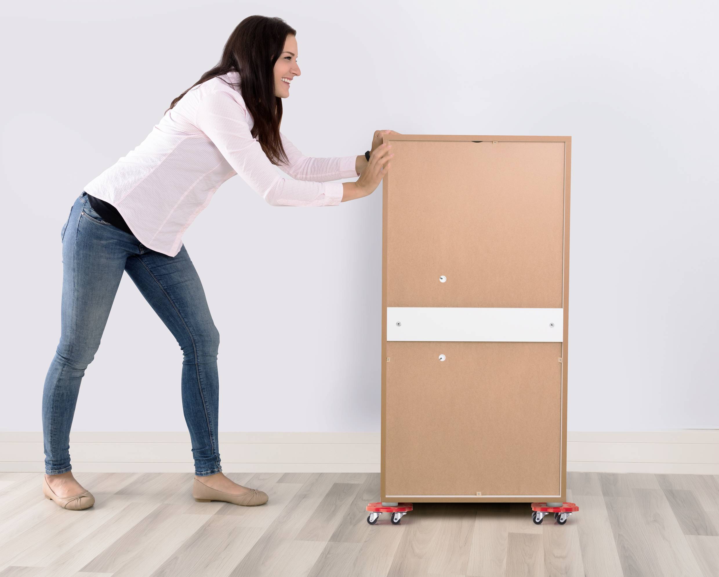 Une femme pousse une armoire en bois sur de petites roulettes à travers un plancher en bois dans une pièce aux murs blancs.