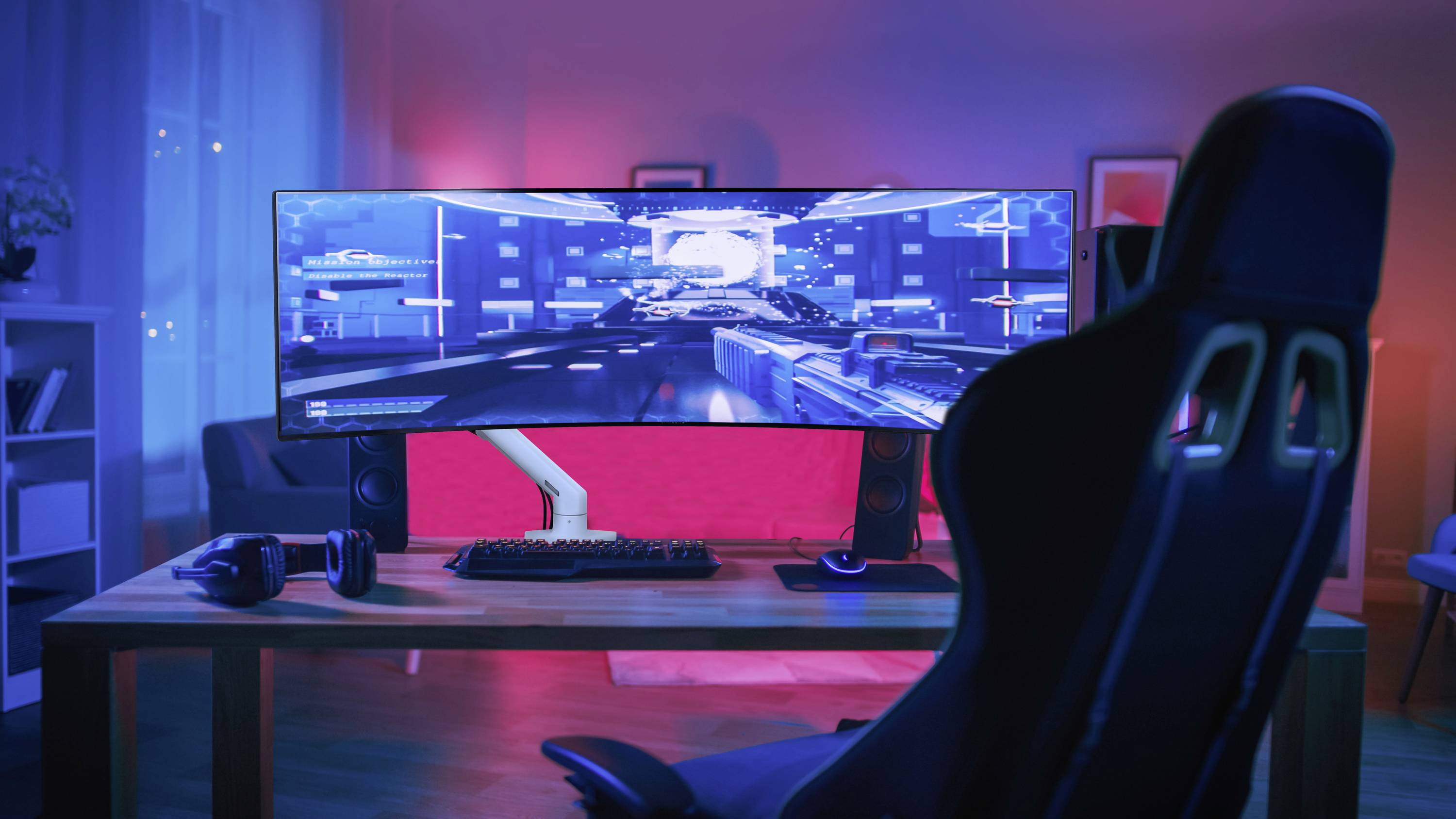 Un bureau informatique avec un grand écran incurvé affiche un jeu vidéo. Une chaise de gaming est placée devant dans une pièce éclairée en rouge et bleu.