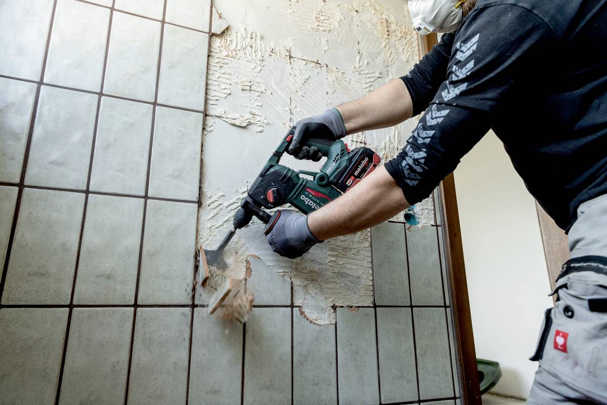 Une personne retire des carreaux d'un mur à l'aide d'un outil électrique, portant un masque de protection et des gants.