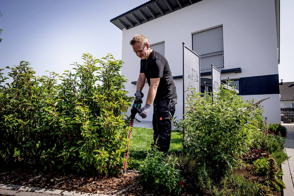 Une personne taille des haies dans un jardin bien entretenu devant une maison individuelle moderne. Le ciel est clair et ensoleillé.