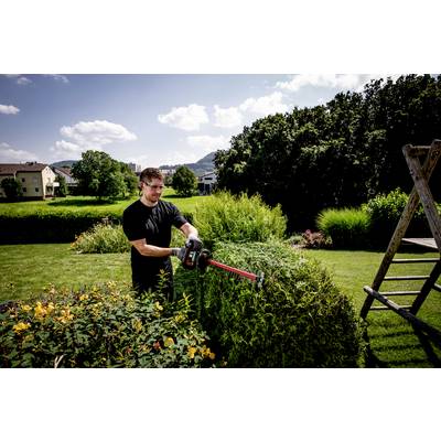 Un homme taille des haies dans un jardin avec un taille-haie électrique. Des arbres et des maisons sont visibles en arrière-plan.