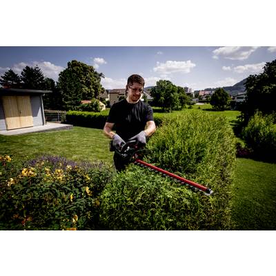 Un homme taille une haie avec un taille-haie électrique dans un jardin bien entretenu. Des maisons sont visibles en arrière-plan.
