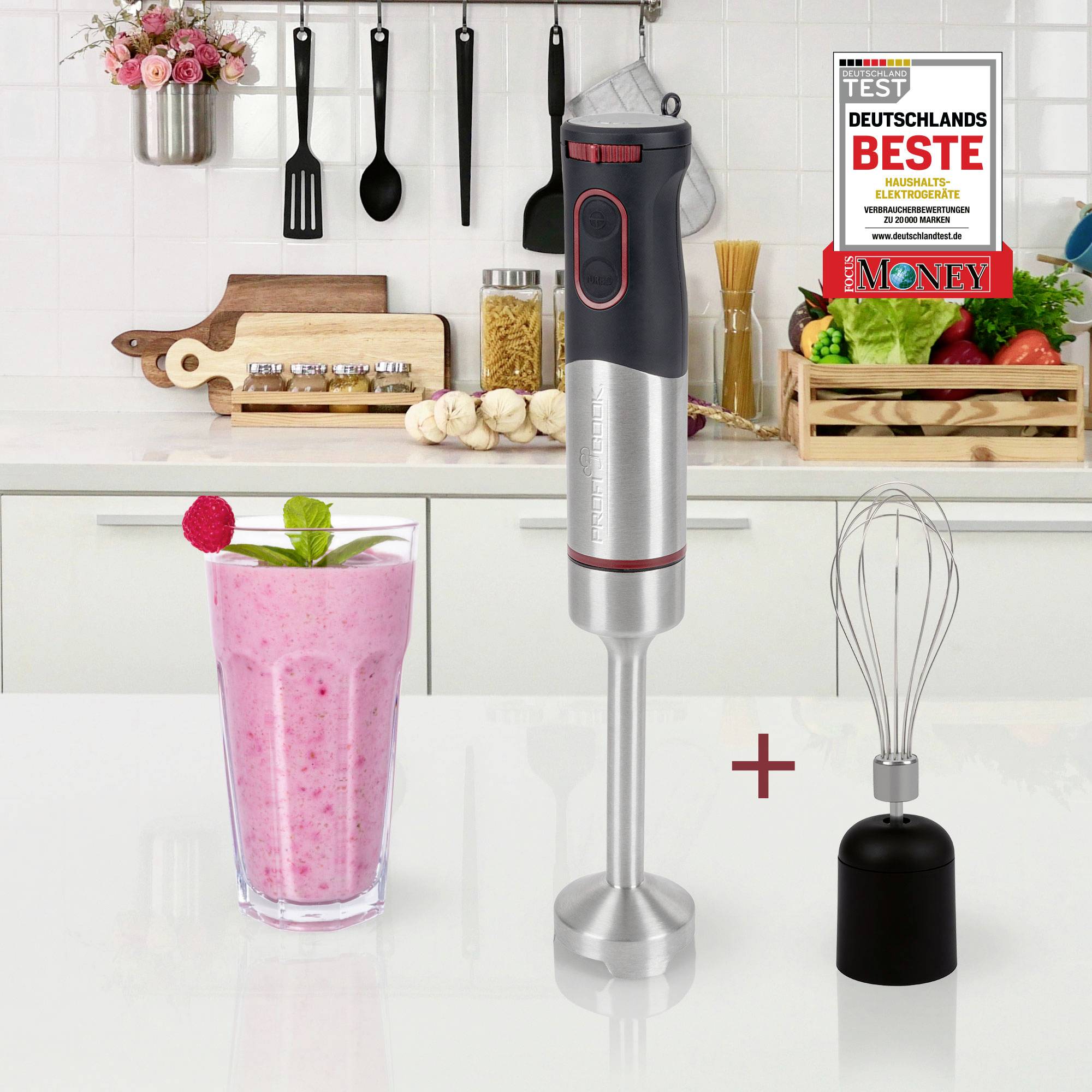 Un mixeur plongeant est posé sur un plan de travail à côté d'un verre de smoothie aux fraises. À l'arrière-plan, un fouet et divers ustensiles de cuisine sont disposés.