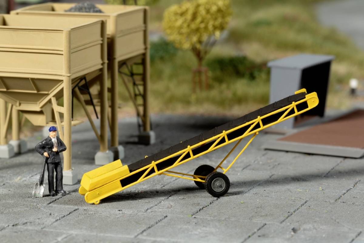 Modèle de jouet d'un chantier de construction avec un ouvrier et un tapis roulant jaune. Des conteneurs de chantier et des arbres miniatures sont visibles en arrière-plan.